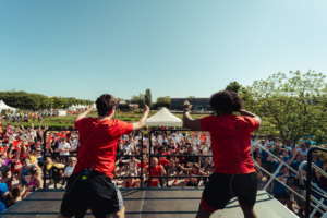 Cette photo contient 2 personnes de dos, debout sur une scène. Equipées de micros serre-tête, elles entrainent la foule devant la scène à un échauffement sportif.